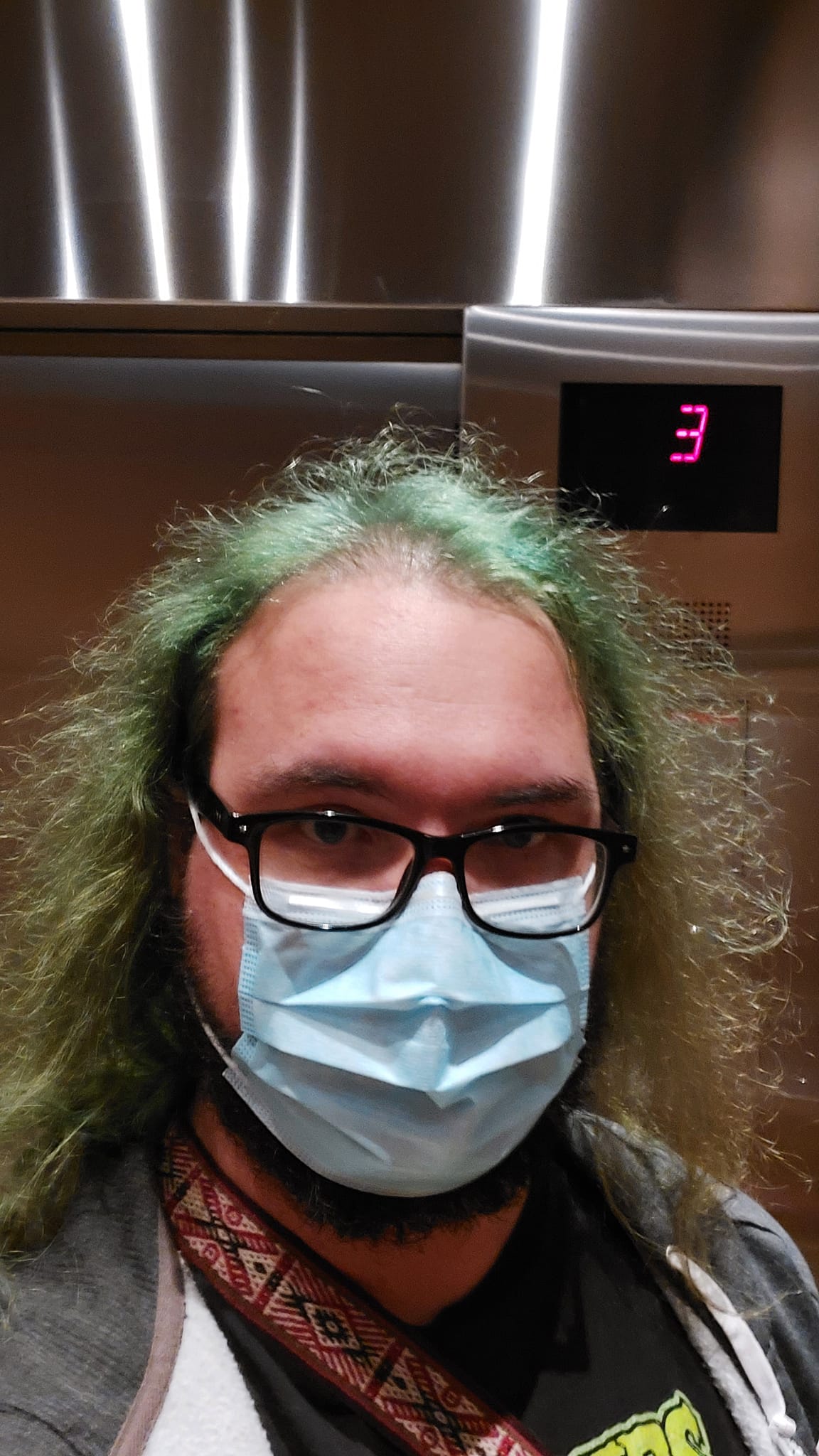 A man with long green hair and a surgical mask stands in an elevator